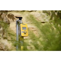 Ein gelbes Bewässerungssteuergerät ist an einem Rohr vor einer beige-gelben Steinwand befestigt, umgeben von unscharfer Vegetation.